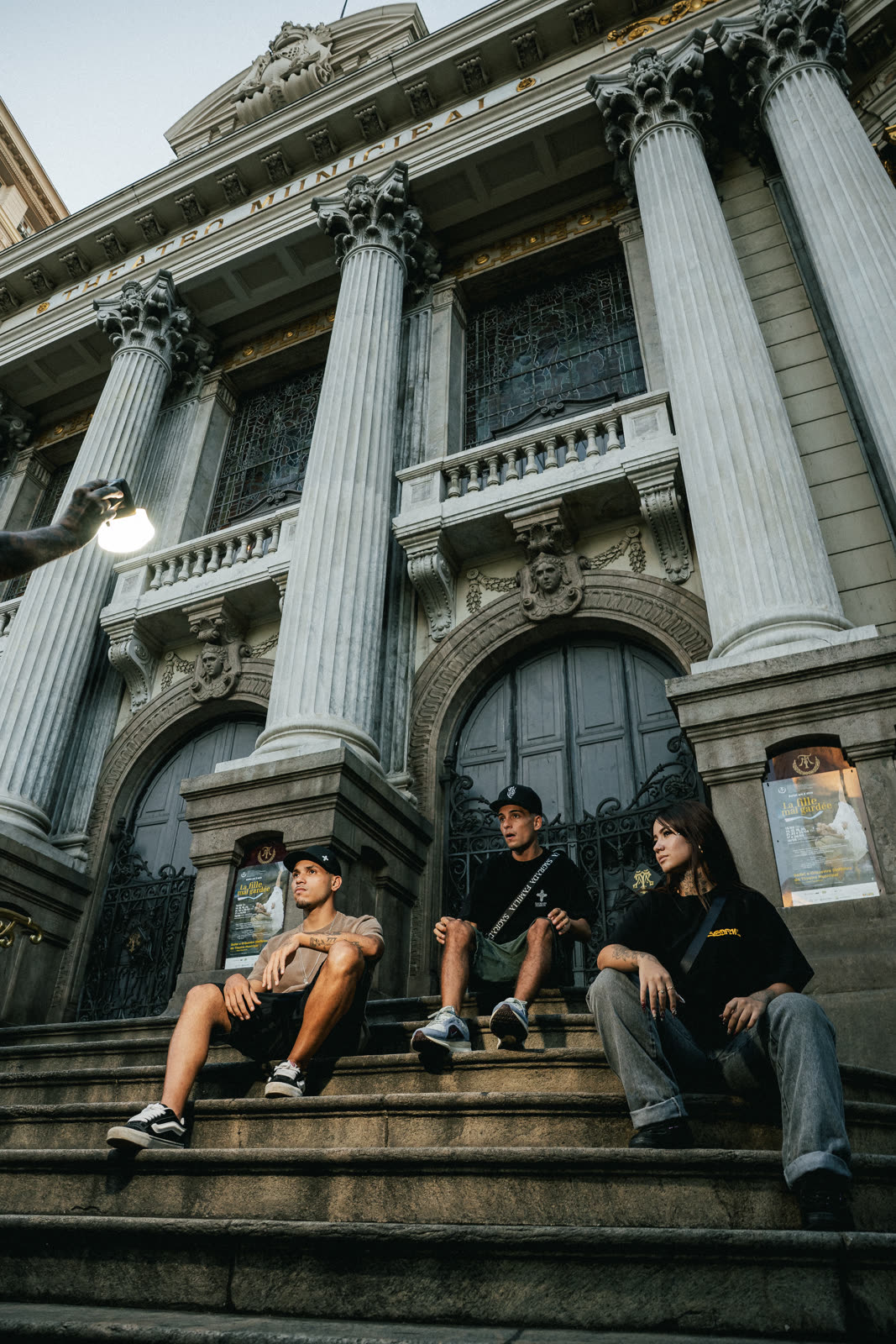 Trio de modelos sentado na escadaria monumental entre colunas coríntias, luz dourada